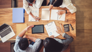 Four people sitting around a table with tablets, computers, and notebooks creating a marketing plan