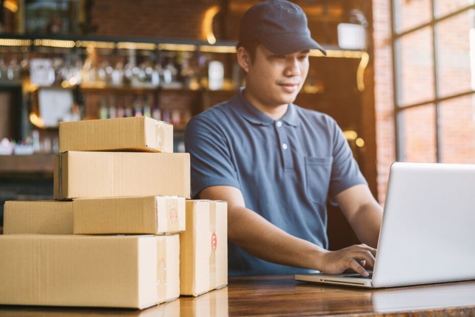 A man in a hat using a computer, next to packages that are ready to ship.